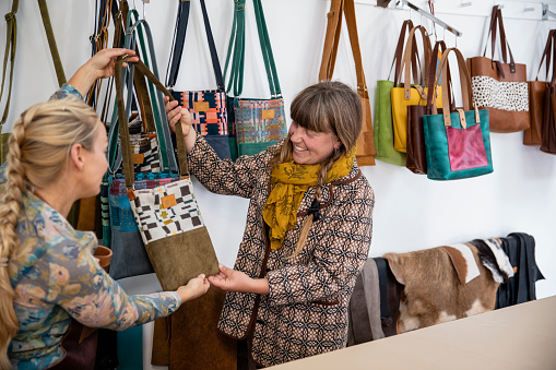A front view of a female business owner showing a customer one of her eco-friendly sustainable leather handbags. She is explaining how all of her products are made from leftover leather and also her commitment to sustainability by using reusable packaging and waste products. / Female Focus Collection