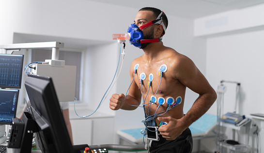 Doctors observing the progress of a cardiopulmonary stress test taken by the male athlete riding a bicycle ergometer. Young adult man having a VO2 test with a VO2 mask on his face, electrocardiogram pads attached, computer recording, indoor bicycle. Healthcare and Medicine concept.