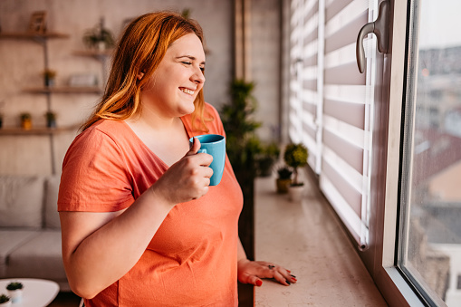Young overweight woman is looking through the window and drinking coffee in the morning.