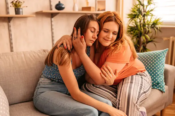 Young woman consoling her sad female friend in living room on sofa.