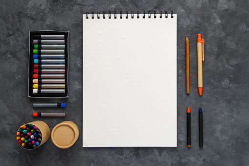 Blank notebook mockup pastel pad dark background flat lay. Minimal work space sketchbook notepad template crayons pencils top view. Back to school banner, stationery, art drawing education e-learning.
