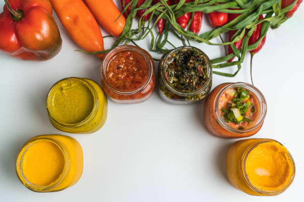 Several glass jars with a variety of traditional latin sauces in a white background Aerial photo of several glass jars with a variety of traditional Latin sauces as huancaina, huacatay, rocoto, chimichurri, anticucho and yellow garlic on a white surface with vegetables. medium group of objects stock pictures, royalty-free photos & images
