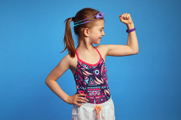 Little girl raises arm and looking at muscles wears goggles and swimsuit Little girl spends leisure time in water park, raises arm and looking at muscles, ready for swim, poses over blue background, wears goggles and swimsuit levitation stock pictures, royalty-free photos & images