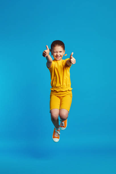 dziewczyna w żółtym stroju skacze, pokazując kciuki w górę na tle niebieskiego - thumbs up child success winning zdjęcia i obrazy z banku zdjęć