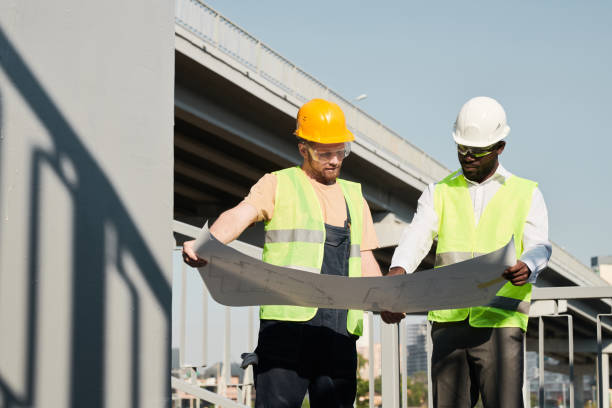 istock Builder in green vest and hardhat analyzing construction plan with African-American manager at site 1337699022