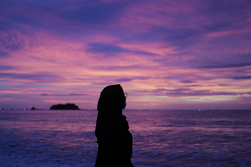 Beautiful young Asian woman in hijab is enjoying a beautiful sunset with the purple and orange sky on the twilight period at Palangpang Beach Sukabumi, West Java, Indonesia.