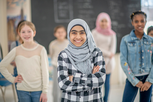 An adorable upper elementary Muslim school girl of Middle Eastern descent is standing in front of her classmates in a classroom. She has her arms crossed over her chest and is smiling widely at the camera.