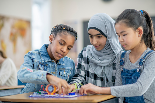 A multiracial group of upper elementary school girls are working together on a circuit board project at a desk in a classroom.