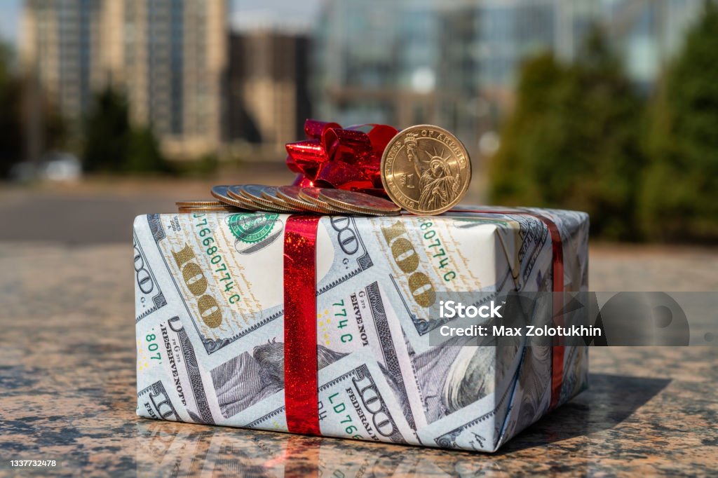 1 American dollar coin, 25 centa coins and a gift wrapped in gift paper depicting 100 American dollars against the background of modern buildings American One Hundred Dollar Bill Stock Photo