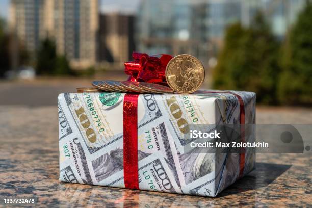 1 American Dollar Coin 25 Centa Coins And A Gift Wrapped In Gift Paper Depicting 100 American Dollars Against The Background Of Modern Buildings Stock Photo - Download Image Now