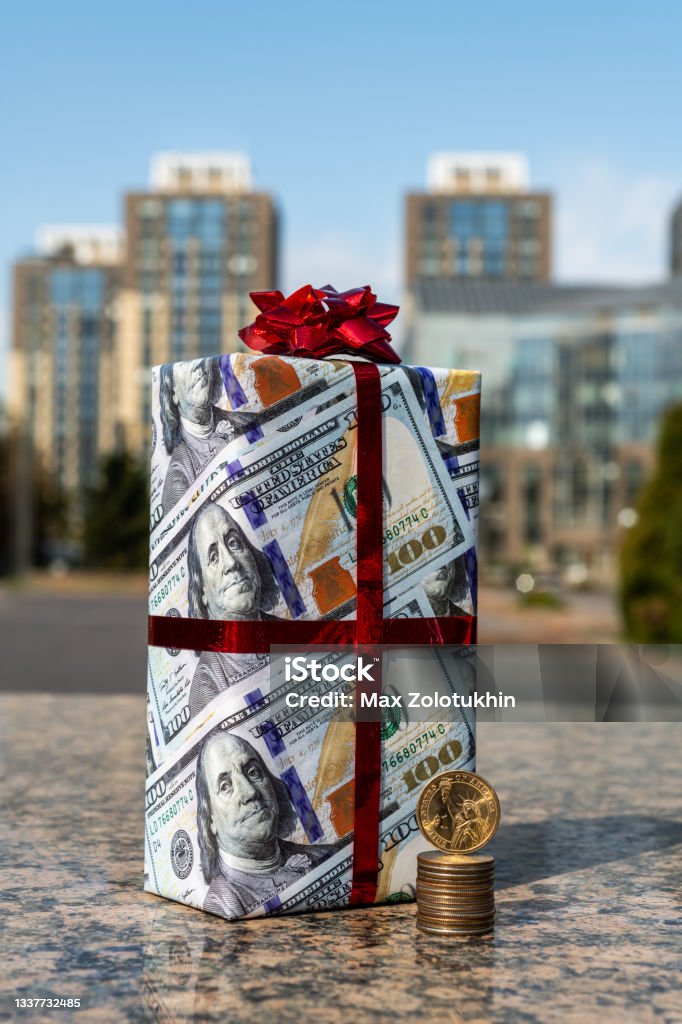 1 American dollar coin, 25 centa coins and a gift wrapped in gift paper depicting 100 American dollars against the background of modern buildings Charitable Donation Stock Photo