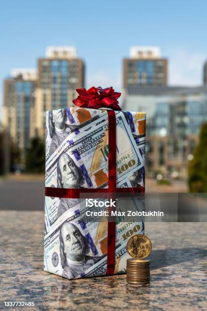 1 American Dollar Coin 25 Centa Coins And A Gift Wrapped In Gift Paper Depicting 100 American Dollars Against The Background Of Modern Buildings Stock Photo - Download Image Now