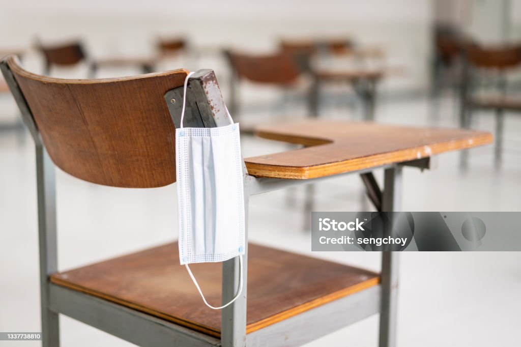 A used medical facemask hangs on a wood lecture chair in the empty classroom during the COVID-19 pandemic School Building Stock Photo