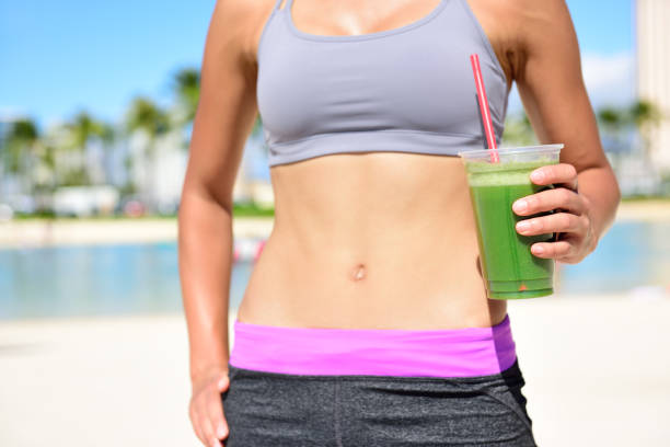 Fitness woman drinking green vegetable smoothie after running exercise. Close up of smoothie and stomach. Healthy lifestyle concept with fit female model outside on beach.