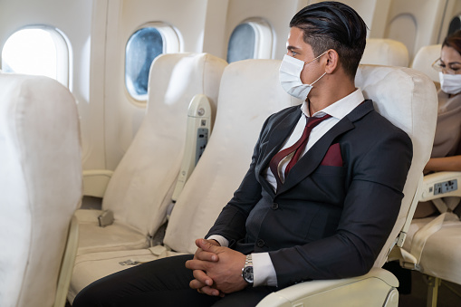 Confident Businessman in formal suit wearing face mask and sitting at seat near window in an airplane. travel and work abroad