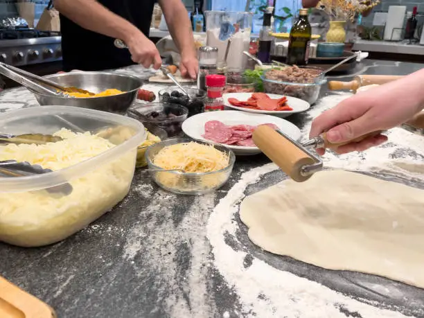 granite countertop covered with flour. Dough being rolled out. Bowls of cheese, salami, pepperoni, olives and other pizza toppings