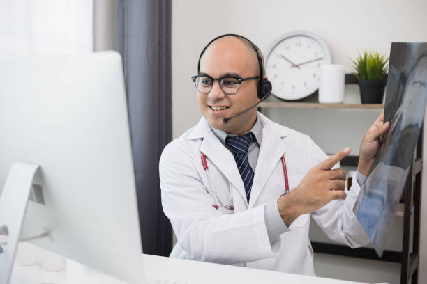 istock Doctor talks to patient via online video conferencing with computer wearing headset. Explain about taking medications to treat illnesses holding x-ray film. 1337752568