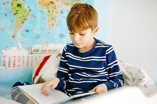 Cute blond little kid boy in pajamas reading book in his bedroom. Excited healthy child reading loud, sitting in his bed. Schoolkid, family, education.