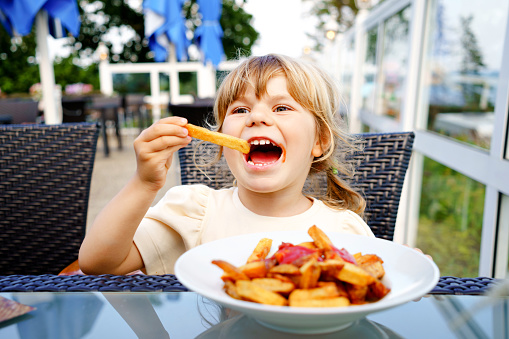 Portrait of happy smiling preschool girl eating french fries with tomato ketchup in restaurant on terrace outdoors. Little child with blond hairs enjoy unhealthy fast food or fresh prepared lunch