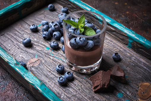 Tasty chocolate pudding or sweet mousse with blueberry and mint in portion glass on wooden tray and dark concrete background