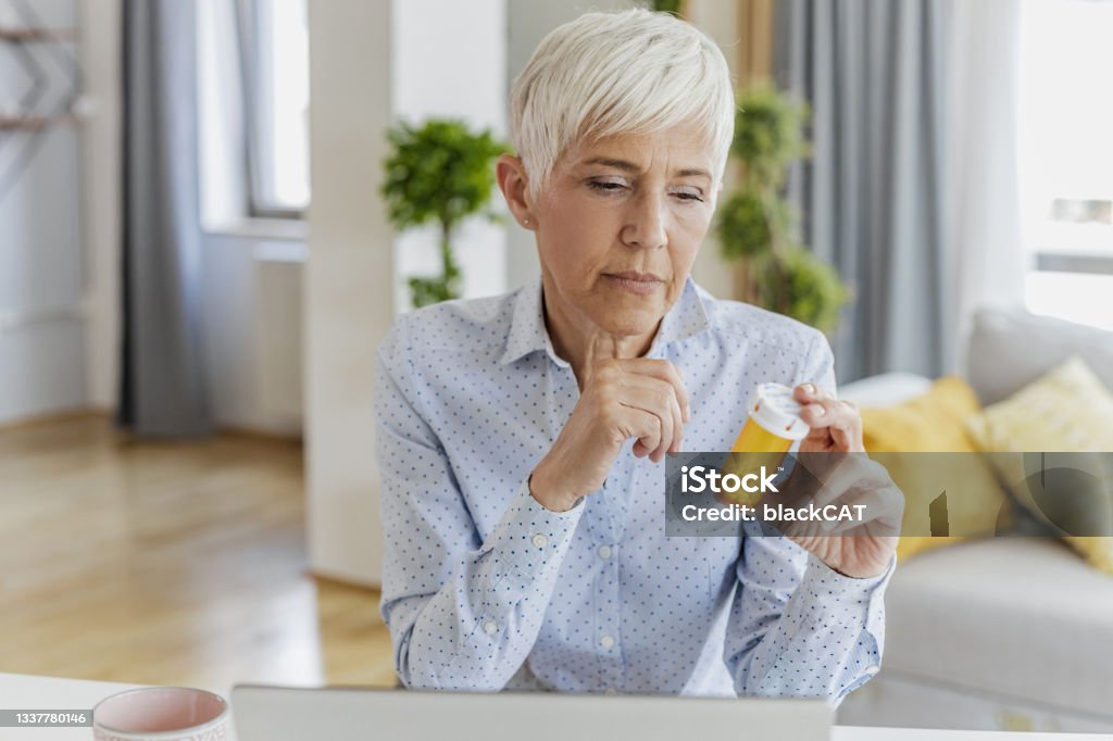 Worried senior woman holding a bottle of medicine in her hand Senior woman sitting at the table in the living room and holding bottle od pills in her hand Taking Medicine Stock Photo
