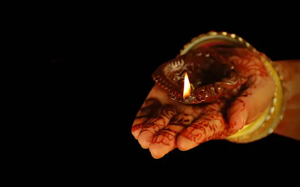 Photo of Diwali, Teej and Karwachauth celebrations themed festive picture of one hand with bangles in hands and mehandi on palms holding a deepak or diya isolated over black