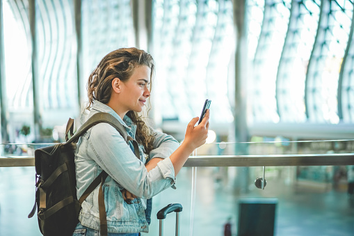 A beautiful caucasian young woman using mobile phone at airport to buy tickets online, book hotel, or rent a car. Covid time travels, space for text