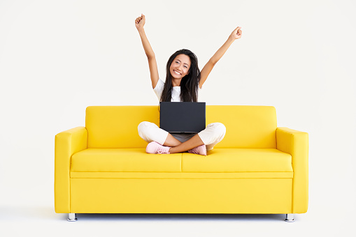 Happy smiling woman with raised arms sitting on yellow couch with laptop and looking to camera on white background. Winning, celebration, success concept