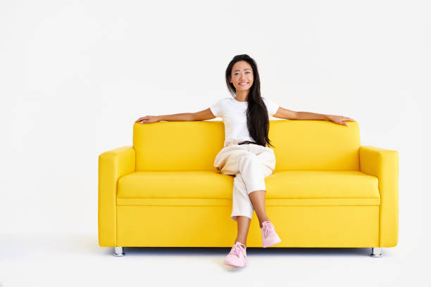 istock Pretty smiling asian female relax on yellow couch on white background 1337793389