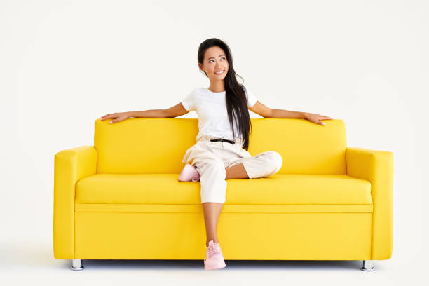 istock Pretty smiling asian female relax on yellow couch on white background 1337793516