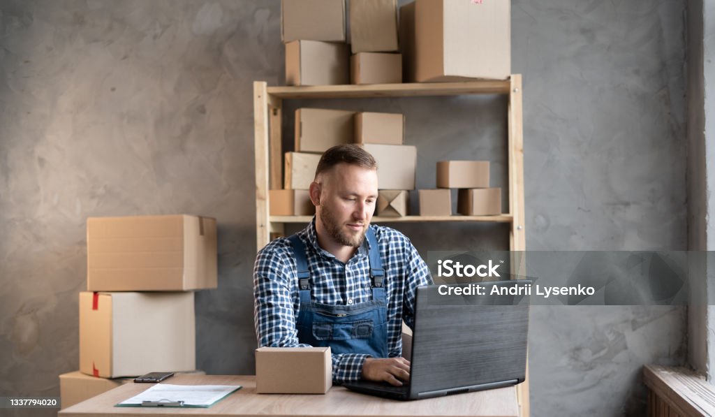 Caucasian male business owner sitting at home office wearing overalls. Caucasian male business owner sitting at home office wearing overalls. using computer, online marketing packaging box delivery, SME e-commerce telemarketing concept. Owner Stock Photo