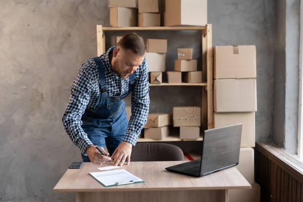 A Caucasian man in a blue overalls prepares goods for shipment, makes notes with a pen. A Caucasian man in a blue overalls prepares goods for shipment, makes notes with a pen. Online sales with start-up and small business, conceptual work from home and online shopping with e-commerce business markup stock pictures, royalty-free photos & images