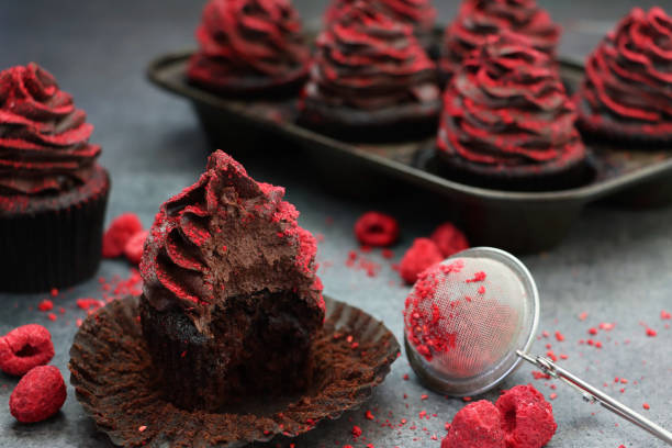 istock Close-up image of individual cupcake with missing bite besides muffin cake tin containing homemade, chocolate cupcakes in brown, paper cake case, chocolate butter icing piped swirl coated with dehydrated raspberry dust, focus on foreground 1337794624