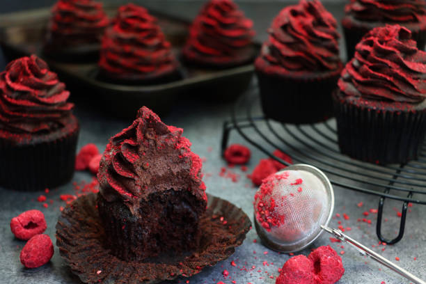 istock Close-up image of individual cupcake with missing bite besides circular, black, metal wire cooling rack containing homemade, chocolate cupcakes in brown, paper cake case, chocolate butter icing piped swirl, dehydrated raspberry dust, focus on foreground 1337794739