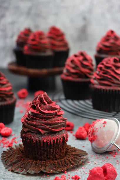 istock Image of individual, unwrapped cupcake besides circular, black, metal wire cooling rack containing homemade, chocolate cupcakes in brown, paper cake case, chocolate butter icing piped swirl, dehydrated raspberry dust, focus on foreground 1337795678