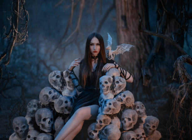 una hermosa y sexy mujer vampiro se sienta en un trono de los cráneos de sus víctimas en un oscuro bosque místico. imagen gótica de la diosa de la muerte con dragón blanco. reina oscura en la colina de los esqueletos - skull gothic style evil dark fotografías e imágenes de stock