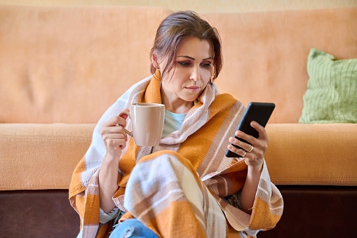 Autumn winter portrait of middle aged woman under warm woolen blanket with hot cup of tea and smartphone in hands. Female resting at home, sitting on the floor near the sofa