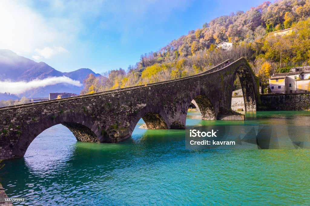 The old name is Devil's Bridge The old name of the bridge is Devil's Bridge. Italy, Lucca. The emerald cold water of the river reflects the ancient asymmetrical arches of the bridge. The Serchio River. Lucca Stock Photo