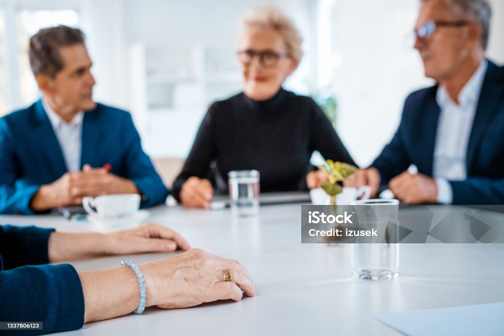 Senior business people during meeting Senior businesswomen and businessmen wearing elegant suits sitting at the table in the office and discussing during business meeting. Close up of hands in the foreground. Business Meeting Stock Photo