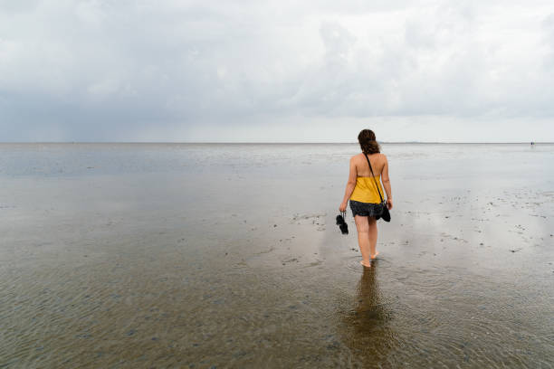 Young woman walking alone in a calm and tranquil beach at low tide in Wadden Sea, Germany. View from behind