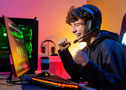 Young teenager plays the computer and celebrates victory in video game with a clenched fist and a smile in room with colored lights.