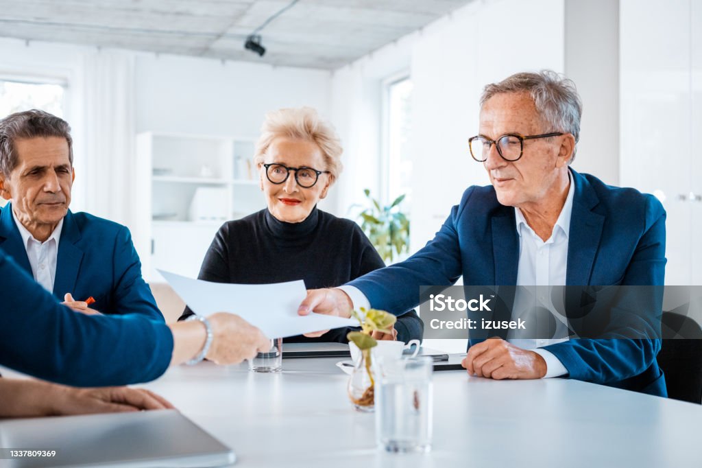 Senior business people during meeting Senior businesswoman and businessmen wearing elegant suits sitting at the table in the office and discussing during business meeting, exchanging contract. Business Stock Photo