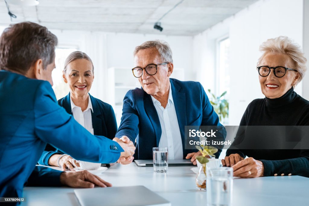 Senior businessmen shaking hands Senior businesswoman and businessmen wearing elegant suits sitting at the table in the office and discussing during business meeting, shaking hands. Business Stock Photo