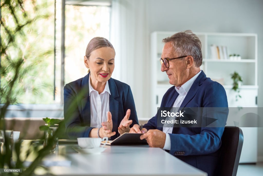 Two senior entrepreneurs in the office Senior businesswoman and businessman wearing elegant suits sitting at the table in the office, working on laptop and discussing. Business Stock Photo