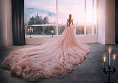 Artistic processing Fantasy girl princess in pink dress stands in medieval room looking vintage window with winter nature landscape mountains sunset. silhouette woman queen long train skirt. Back view