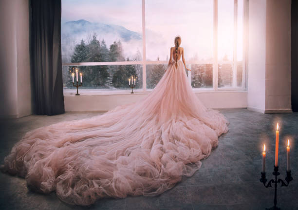 artistic processing fantasy girl princess in pink dress stands in medieval room looking vintage window with winter nature landscape mountains sunset. silhouette woman queen long train skirt. back view - evening gown imagens e fotografias de stock