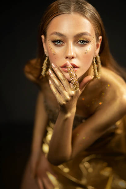 junge modelfrau mit goldenem körper posiert im studio. der hintergrund ist schwarz. perfektes make-up gold, glitzer. mädchen glamouröse königin. schmuckring, ohrringe, accessoires elegantes kleid. schönes gesicht - painted eyes stock-fotos und bilder