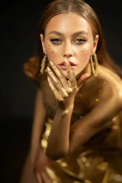 Photo of Young fashion model woman with golden body posing in studio. Background is black. Perfect makeup gold, glitter. Girl glamorous queen. Jewelry ring, earrings, accessories elegant dress. Beautiful face