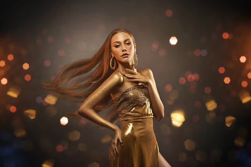 Young stylish girl fashion model in gold dress, outfit posing in studio. Long blond hair flies in wind motion. Beautiful woman with golden skin, body in paint. Background glows, glitters, yellow bokeh