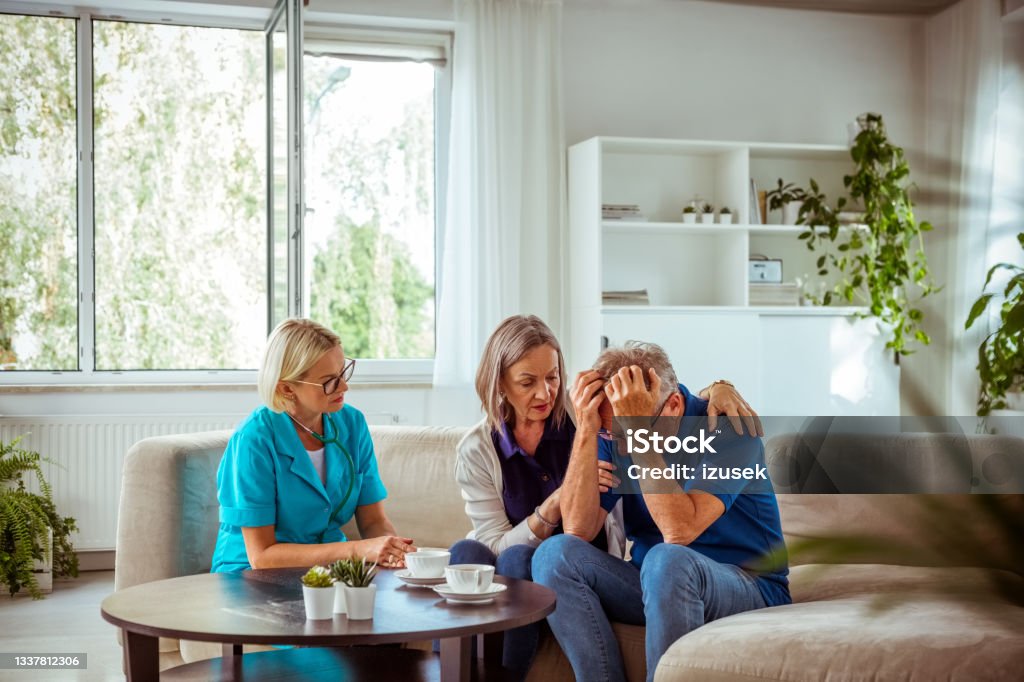 Nurse talking with worried senior couple Worried senior man feeling unwell, getting bad news. Senior woman consoling her husband. They are sitting with nurse in hospital waiting room. Head In Hands Stock Photo